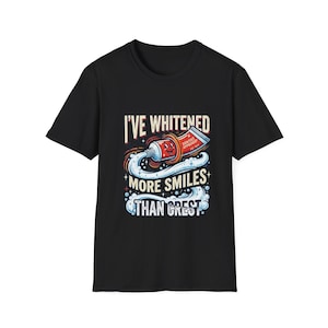 Op de afbeelding: Een zwart T-shirt met korte mouwen en een afbeelding van een rode tandpastatube met een lachend gezicht. De tekst luidt I'VE WHITENED MORE SMILES THAN CREST, met DENTIST APPROVED EST. 1978 op de tube, omringd door witte tandpastawervelingen.