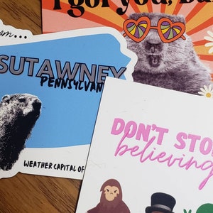 May include: A collection of colorful stickers and postcards. One features a groundhog wearing heart-shaped sunglasses with the text "I got you, babe." Another says "Punxsutawney, Pennsylvania" and "Weather Capital of." A third says "Don't stop believing."