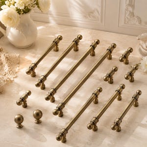 May include: A collection of antique brass cabinet hardware including several long bar pulls with spherical ends, smaller T-shaped pulls, and two round knobs. The metal hardware features a brushed, weathered finish with visible cylindrical details.