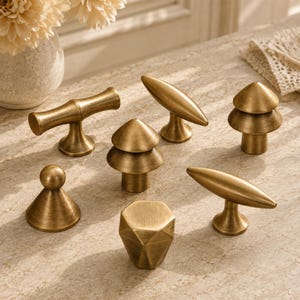 May include: A collection of seven brass cabinet knobs and pulls in various geometric and organic shapes, including conical, cylindrical, and faceted designs. Each piece features a brushed metallic finish with a warm, golden-toned surface.