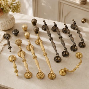May include: A collection of metal cabinet hardware including various drawer pulls and knobs in brushed gold and antique bronze finishes. The set features elongated handles with ornate detailing, circular base plates, and round textured knobs.