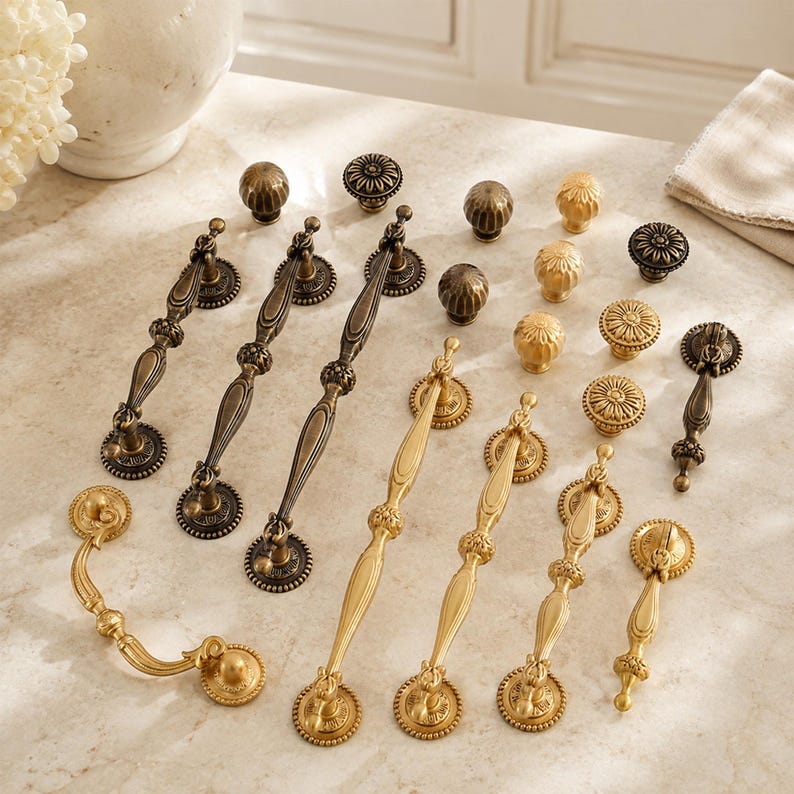 May include: A collection of metal cabinet hardware including various drawer pulls and knobs in antique bronze and brushed gold finishes. The assortment features elongated handles with ornate detailing and circular knobs with textured patterns.