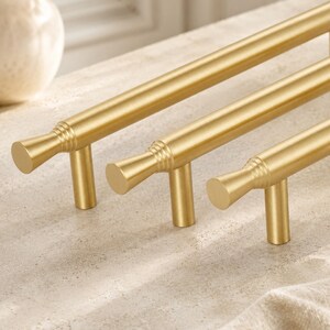 May include: Three brushed gold cabinet pulls arranged in a row. Each handle features a long, cylindrical bar with a flared, circular end cap detailed with three horizontal grooves where the bar meets the mounting post and the end piece.