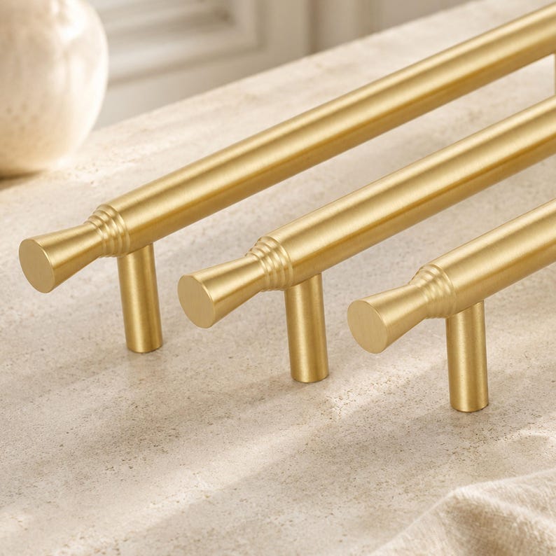 May include: Three brushed gold cabinet pulls arranged in a row. Each handle features a long, cylindrical bar with a flared, circular end cap detailed with three horizontal grooves where the bar meets the mounting post and the end piece.