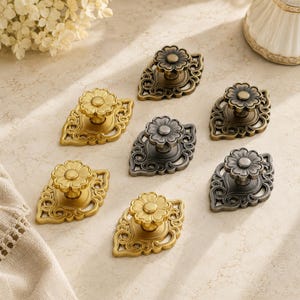 Puede incluir: Un conjunto de seis pomos de metal para cajones, cada uno con un tirador central en forma de flor montado sobre una placa posterior ornamentada con forma de hoja. El set incluye tres pomos en acabado dorado pulido y tres en bronce antiguo.