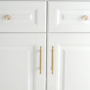May include: Two white cabinet drawers featuring round, gold-toned button-style knobs, positioned above two white cabinet doors equipped with vertical, cylindrical gold-toned bar handles. The hardware provides a metallic contrast to the white cabinetry.