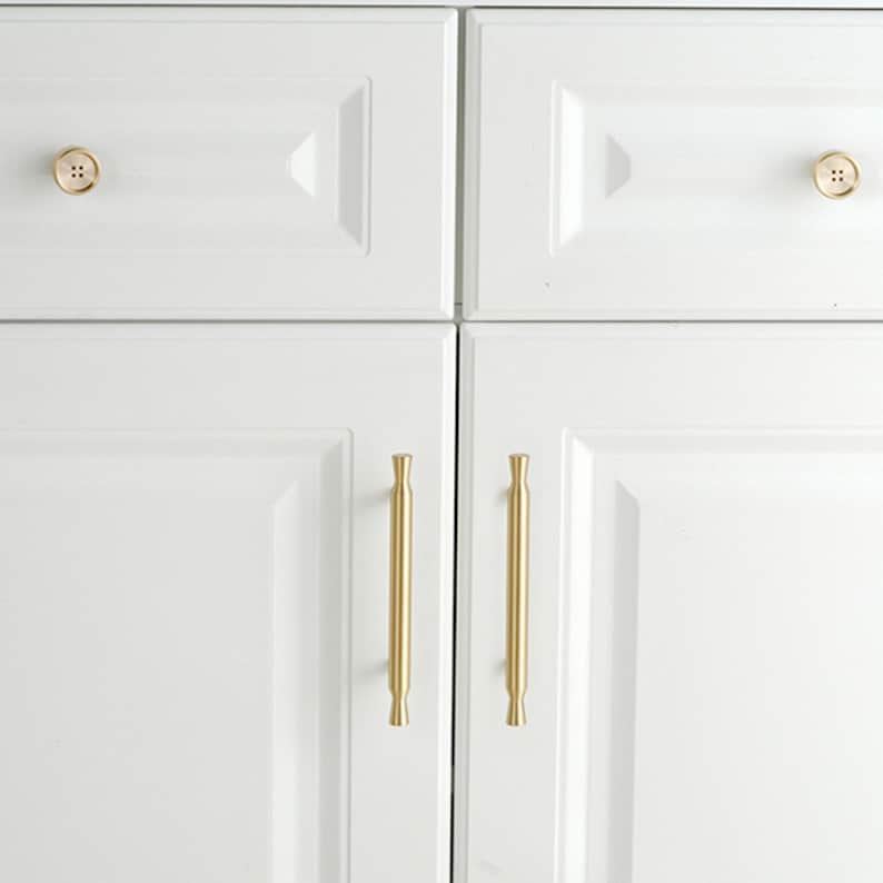 May include: Two white cabinet drawers featuring round, gold-toned button-style knobs, positioned above two white cabinet doors equipped with vertical, cylindrical gold-toned bar handles. The hardware provides a metallic contrast to the white cabinetry.