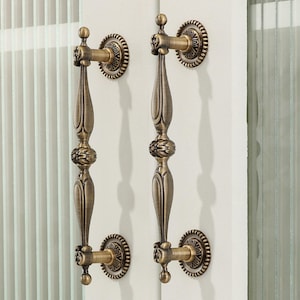 May include: Two vertical door handles with an antique brass finish, featuring a central decorative floral motif and elongated, tapered shafts. Each handle is secured by two circular mounting plates with a beaded edge design on a white door.