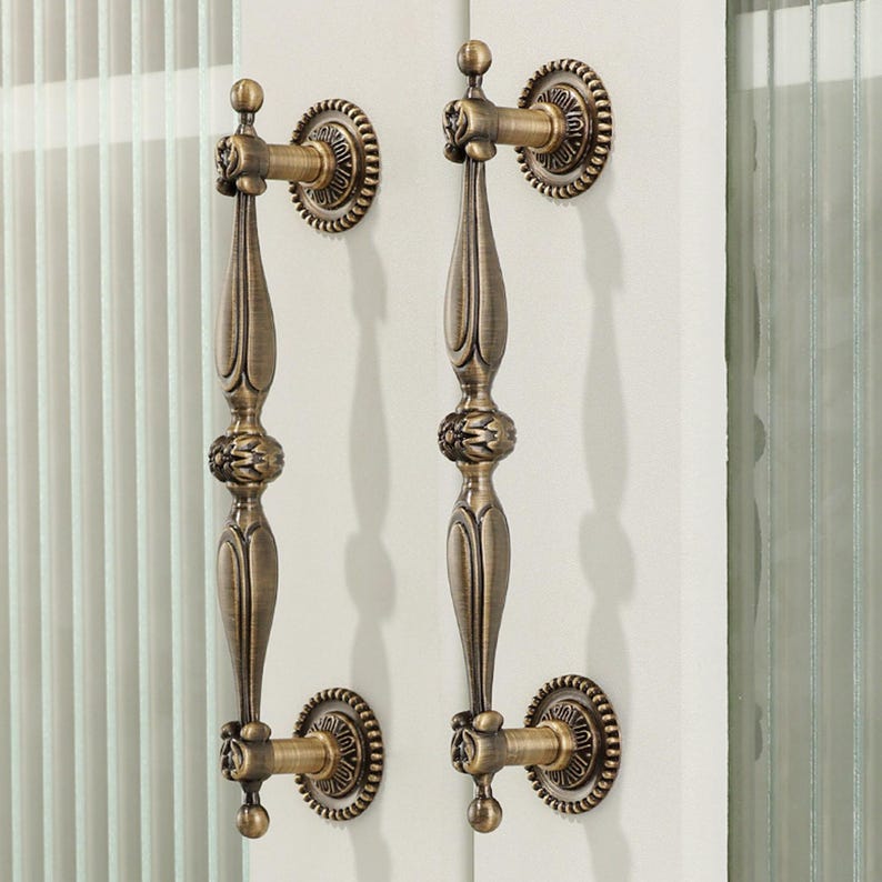 May include: Two vertical door handles with an antique brass finish, featuring a central decorative floral motif and elongated, tapered shafts. Each handle is secured by two circular mounting plates with a beaded edge design on a white door.