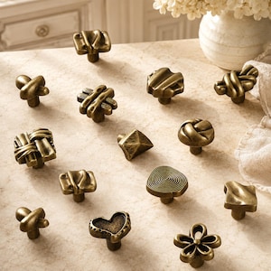 Könnte beinhalten: Eine Sammlung von dreizehn messingfarbenen Metall-Möbelknöpfen in verschiedenen Formen, darunter geflochtene, geknotete, spiralförmige, herzförmige und florale Designs. Jeder Knopf hat einen zentralen Befestigungsstift und eine texturierte, antik wirkende Metalloberfläche.