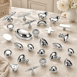 May include: A collection of various polished chrome cabinet knobs and pulls in diverse shapes including round, oval, T-bar, cross, and abstract forms. These metallic hardware pieces feature a reflective, smooth finish and are arranged in a group.