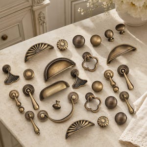 May include: A collection of antique brass cabinet hardware including various drawer pulls, knobs, and drop handles. The assortment features semi-circular cup pulls, round knobs with textured edges, and ornate drop-style handles in a metallic finish.