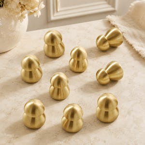 Può includere: Un set di nove piccoli pomelli per armadietti a forma di fungo realizzati in ottone spazzolato. Ogni pomello presenta una parte superiore liscia e arrotondata e una base affusolata, con una finitura oro metallizzato e una texture opaca.