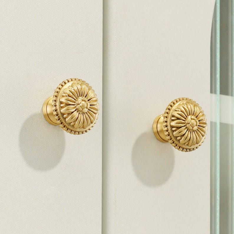 May include: Two circular brass cabinet knobs featuring a raised, detailed floral petal design on the front face. Each knob has a textured, beaded border around the edge and is mounted onto a light-colored, flat vertical surface.