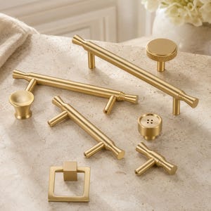 May include: A collection of brushed gold cabinet hardware including two long bar pulls, one medium bar pull, one small T-bar pull, one square drop pull, one round knob with a textured edge, and one round knob featuring four small holes in the center.