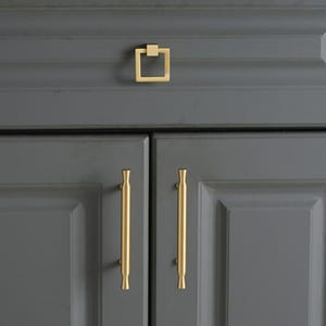 May include: A set of gold-toned cabinet hardware featuring one square-shaped drop pull and two vertical bar handles with flared ends. The hardware is mounted on dark gray cabinet doors and a drawer, showcasing a smooth, metallic finish.