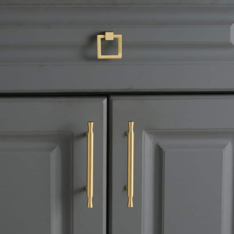 May include: A set of gold-toned cabinet hardware featuring one square-shaped drop pull and two vertical bar handles with flared ends. The hardware is mounted on dark gray cabinet doors and a drawer, showcasing a smooth, metallic finish.