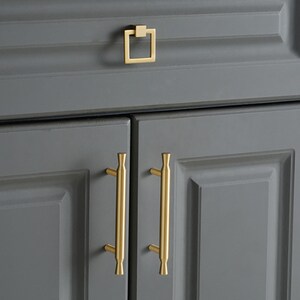 May include: A set of gold-toned cabinet hardware featuring two vertical bar pulls with flared ends and one square drop pull. The hardware is mounted on dark gray cabinet doors and a drawer, showcasing a smooth metallic finish and clean lines.