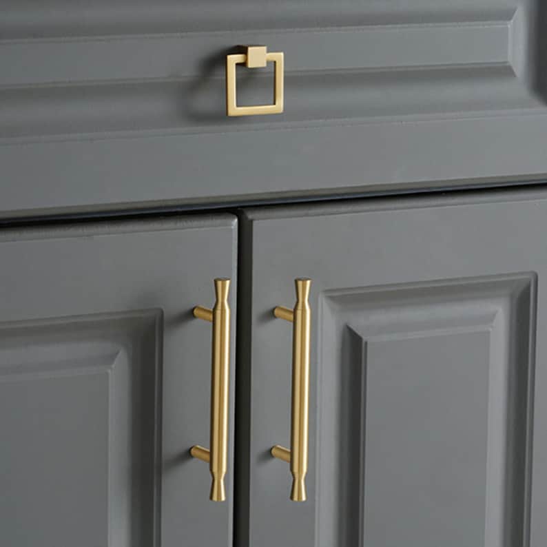 May include: A set of gold-toned cabinet hardware featuring two vertical bar pulls with flared ends and one square drop pull. The hardware is mounted on dark gray cabinet doors and a drawer, showcasing a smooth metallic finish and clean lines.
