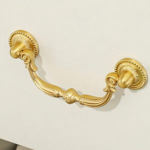 May include: A gold-colored metal drawer pull featuring a curved handle with decorative scrollwork and fluted detailing. The handle is attached to two circular mounting plates, each finished with a beaded edge and a central knob-like protrusion.