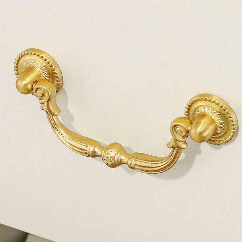 May include: A gold-colored metal drawer pull featuring a curved handle with decorative scrollwork and fluted detailing. The handle is attached to two circular mounting plates, each finished with a beaded edge and a central knob-like protrusion.