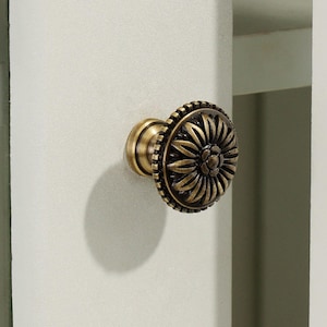 May include: A round cabinet knob with an antique brass finish featuring a raised floral petal design. The circular face displays a central flower surrounded by radiating petals and a textured outer edge, mounted on a light-colored cabinet door.