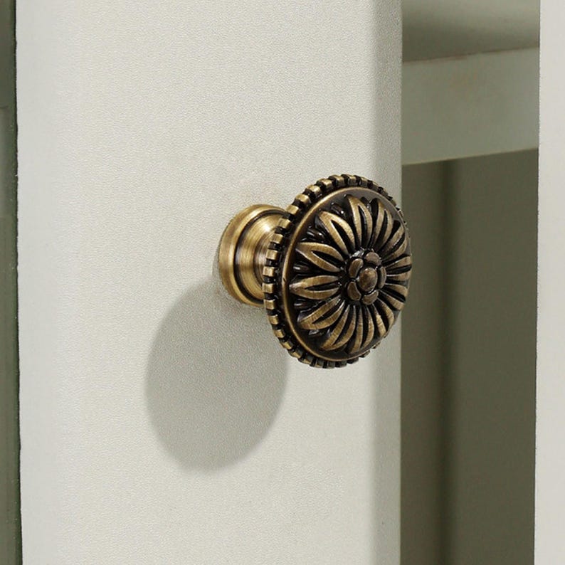 May include: A round cabinet knob with an antique brass finish featuring a raised floral petal design. The circular face displays a central flower surrounded by radiating petals and a textured outer edge, mounted on a light-colored cabinet door.