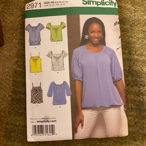 May include: Simplicity sewing pattern 2971 with multiple blouse designs. Sizes include H5: 6, 8, 10, 12, and 14. The image shows a woman wearing a light purple blouse. The pattern also includes a variety of blouse styles, with EUR sizes 32-40 and FR sizes 34-42.