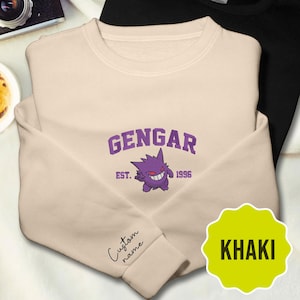 Könnte beinhalten: Ein khakifarbenes Sweatshirt mit Rundhalsausschnitt und einer violetten Stickerei des Charakters Gengar sowie dem Text GENGAR EST. 1996. Der Ärmel zeigt den gestickten Schriftzug Custom name in Schreibschrift, ein gelbes Sternetikett zeigt KHAKI.
