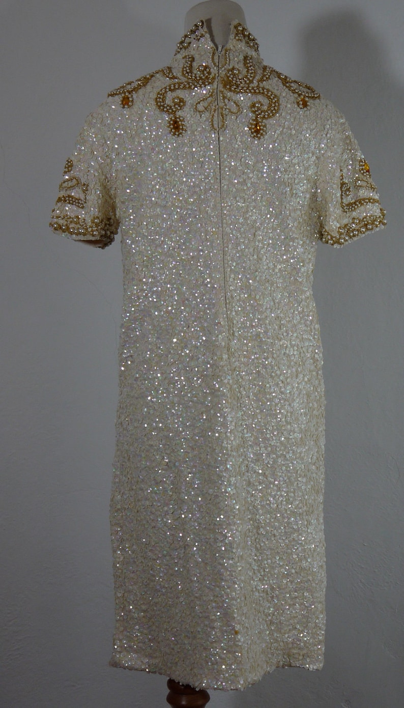 K&ouml;nnte beinhalten: Elfenbeinfarbenes, knielanges Kleid mit Pailletten. Das Kleid hat einen hohen, verzierten Ausschnitt und kurze &Auml;rmel mit Perlenakzenten. Der R&uuml;cken hat einen Rei&szlig;verschluss. Das Kleid wird auf einer Schaufensterpuppe pr&auml;sentiert.