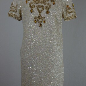 K&ouml;nnte beinhalten: Ein cremefarbenes Paillettenkleid mit kurzen &Auml;rmeln und hohem Ausschnitt. Das Kleid ist mit goldenen und perlenartigen Verzierungen am Ausschnitt, an den &Auml;rmeln und am Oberteil verziert. Das Kleid ist knielang und scheint vintage zu sein.