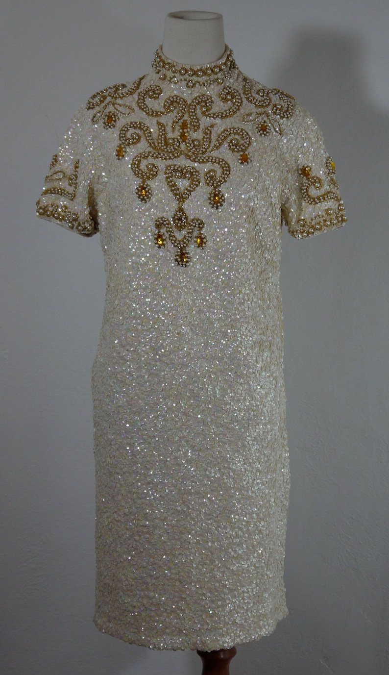 K&ouml;nnte beinhalten: Ein cremefarbenes Paillettenkleid mit kurzen &Auml;rmeln und hohem Ausschnitt. Das Kleid ist mit goldenen und perlenartigen Verzierungen am Ausschnitt, an den &Auml;rmeln und am Oberteil verziert. Das Kleid ist knielang und scheint vintage zu sein.