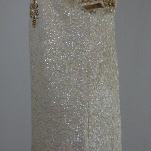 K&ouml;nnte beinhalten: Elfenbeinfarbenes Paillettenkleid mit hohem Ausschnitt und kurzen &Auml;rmeln. Das Kleid ist mit goldfarbenen Perlen und Pailletten am Ausschnitt und an den &Auml;rmeln verziert. Das Kleid wird auf einer Schaufensterpuppe pr&auml;sentiert.