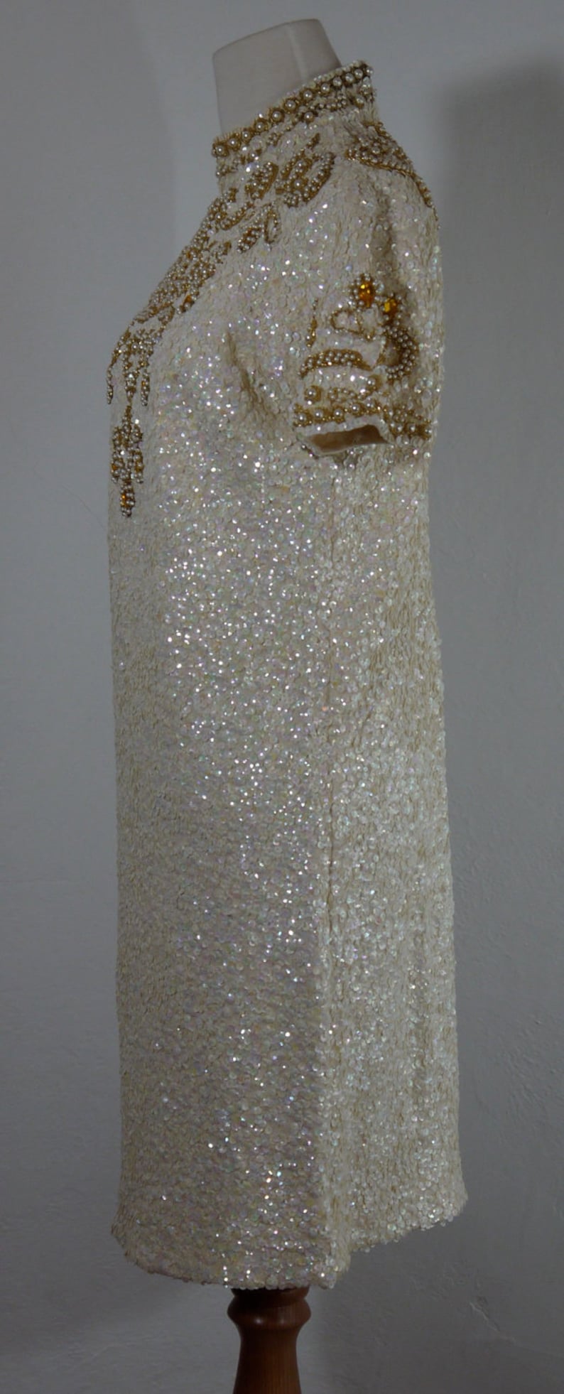 K&ouml;nnte beinhalten: Elfenbeinfarbenes Paillettenkleid mit hohem Ausschnitt und kurzen &Auml;rmeln. Das Kleid ist mit goldfarbenen Perlen und Pailletten am Ausschnitt und an den &Auml;rmeln verziert. Das Kleid wird auf einer Schaufensterpuppe pr&auml;sentiert.