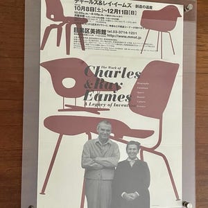 Pode incluir: Cartaz emoldurado com a obra de Charles & Ray Eames. O cartaz, em tons de bordô e branco, mostra desenhos de cadeiras e retratos. O texto inclui detalhes da exposição. A moldura é de vidro.