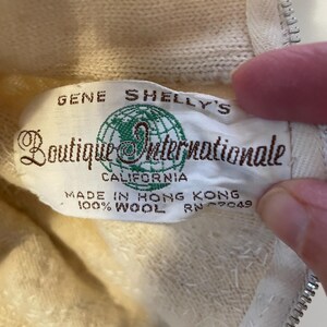 K&ouml;nnte beinhalten: Nahaufnahme eines Vintage-Etiketts von "Gene Shelly's Boutique Internationale" in Creme und Gr&uuml;n. Auf dem Etikett steht "Made in Hong Kong, 100% Wool". Das Kleidungsst&uuml;ck ist hellcremefarben mit einem Rei&szlig;verschluss.
