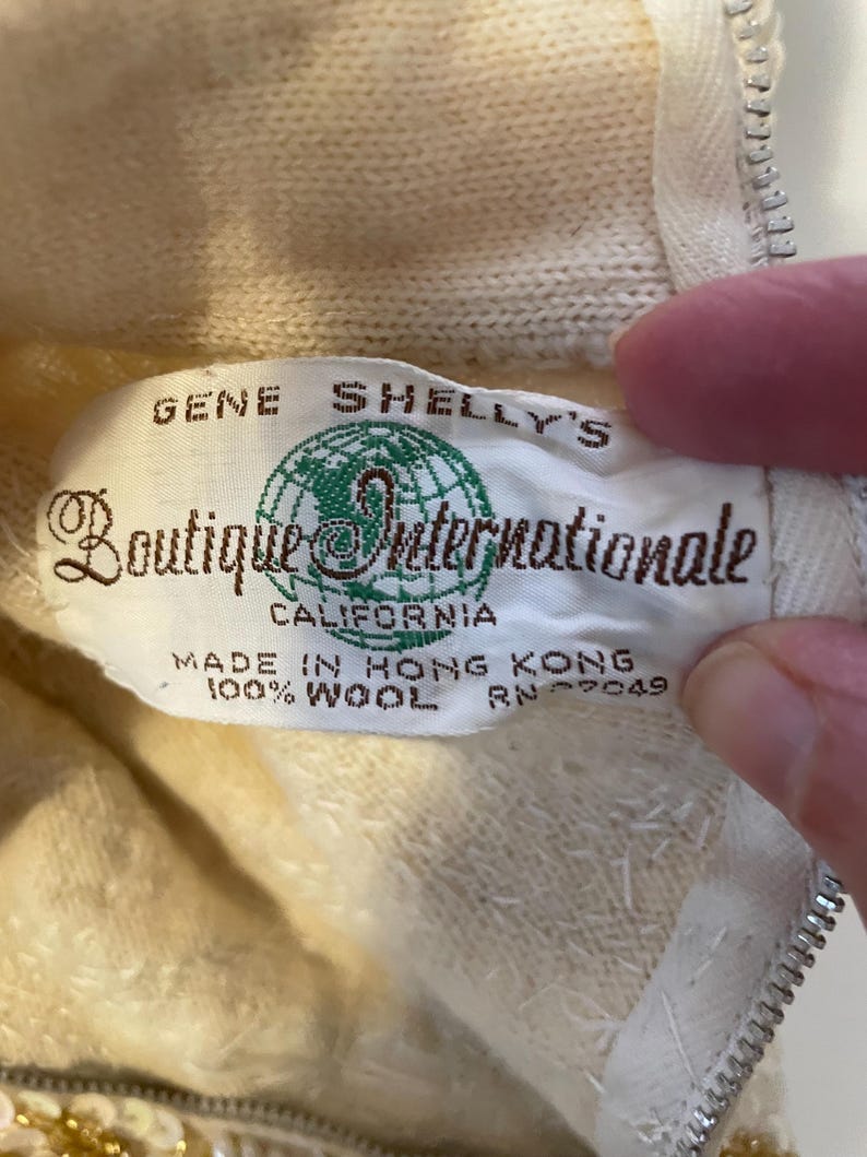 K&ouml;nnte beinhalten: Nahaufnahme eines Vintage-Etiketts von "Gene Shelly's Boutique Internationale" in Creme und Gr&uuml;n. Auf dem Etikett steht "Made in Hong Kong, 100% Wool". Das Kleidungsst&uuml;ck ist hellcremefarben mit einem Rei&szlig;verschluss.
