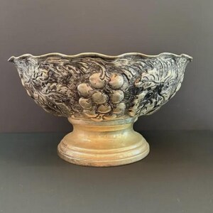 May include: A silver-toned metal pedestal bowl featuring a scalloped rim and a detailed embossed design of grape clusters and vine leaves wrapping around the exterior. The bowl stands on a circular base with a smooth, polished finish.