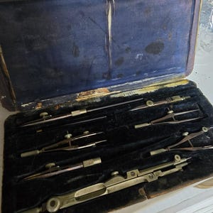 May include: A vintage drafting instrument set housed in a rectangular case with a dark blue velvet interior. The set includes several brass and metal compasses and dividers with adjustable screws, arranged in custom-fitted slots within the case.