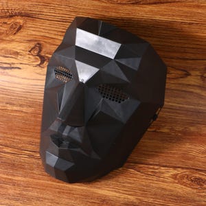 Può includere: Una maschera nera a copertura totale del viso con un design geometrico sfaccettato e piani angolari netti. La maschera presenta aperture a rete integrate per gli occhi e una struttura modellata per naso, bocca e mento.