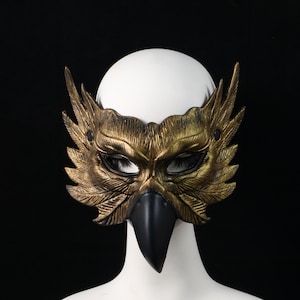 Könnte beinhalten: Eine goldfarbene Vogelmaske mit detaillierten Federstrukturen und einem markanten schwarzen Schnabel. Die Maske bedeckt den Augenbereich und erstreckt sich mit flügelartigen Formen zu beiden Seiten, befestigt mit kleinen schwarzen Nieten.