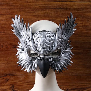 Könnte beinhalten: Eine silber-schwarze Eulenmaske mit detaillierten Federstrukturen, markanten spitzen Federohren und einem glatten schwarzen Schnabel. Die Maske wird auf einem weißen Schaufensterpuppenkopf präsentiert und zeigt Augenöffnungen mit Wimpern.
