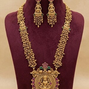 May include: A gold-toned necklace and earring set featuring intricate deity motifs, small beaded chains, and accents of pink and green stones. The large central pendant displays a seated figure surrounded by a circular filigree frame and dangling beads.