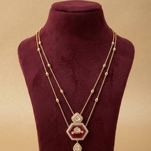 Puede incluir: Un collar de doble cadena de oro con un colgante hexagonal central con incrustaciones de piedra roja oscura y pequeñas piedras claras. El colgante incluye un dije en forma de lágrima y la cadena está adornada con pequeñas cuentas doradas.