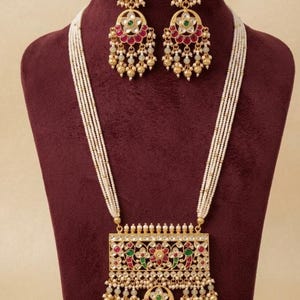 Puede incluir: Un conjunto de collar y pendientes en tono dorado con múltiples hilos de pequeñas cuentas blancas. El colgante rectangular y los pendientes a juego están decorados con piedras rojas y verdes, cristales transparentes y cuentas colgantes.