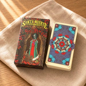 May include: A Santa Muerte Tarot deck featuring a box with a skeletal figure in a red and green robe surrounded by red roses and the text "SANTA MUERTE TAROT BOOK OF THE DEAD". Beside it, a stack of cards displays a circular pattern of skulls and flowers.