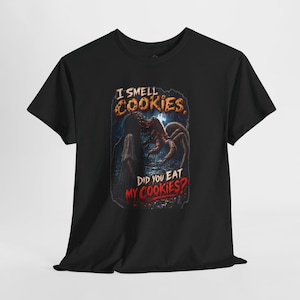 May include: A black short-sleeve t-shirt featuring a graphic print of a dark, monstrous creature with long limbs and a figure in a white dress. The text reads I SMELL COOKIES and DID YOU EAT MY COOKIES? in a distressed, horror-themed font style.