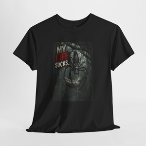 May include: A black short-sleeved t-shirt featuring a graphic print of a large spider hanging from a web in a dark forest. The text "MY LIFE SUCKS..." is printed in white and red lettering to the left of the spider, which is holding a cocoon.