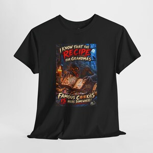 May include: A black t-shirt featuring a graphic print of a creature reading a book titled Grandma's Recipes. The text reads: I KNOW THAT THE RECIPE FOR GRANDMA'S FAMOUS COOKIES IS HERE SOMEWHERE! The scene includes a candle, cookies, and a window.
