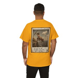 May include: A mustard yellow short-sleeve t-shirt featuring a rectangular graphic on the back. The print depicts a vintage-style photograph of a person in a red wagon flying over a canyon, with the text "April 4, 1969" and "As you can see Grandma was a bit wild in her day."